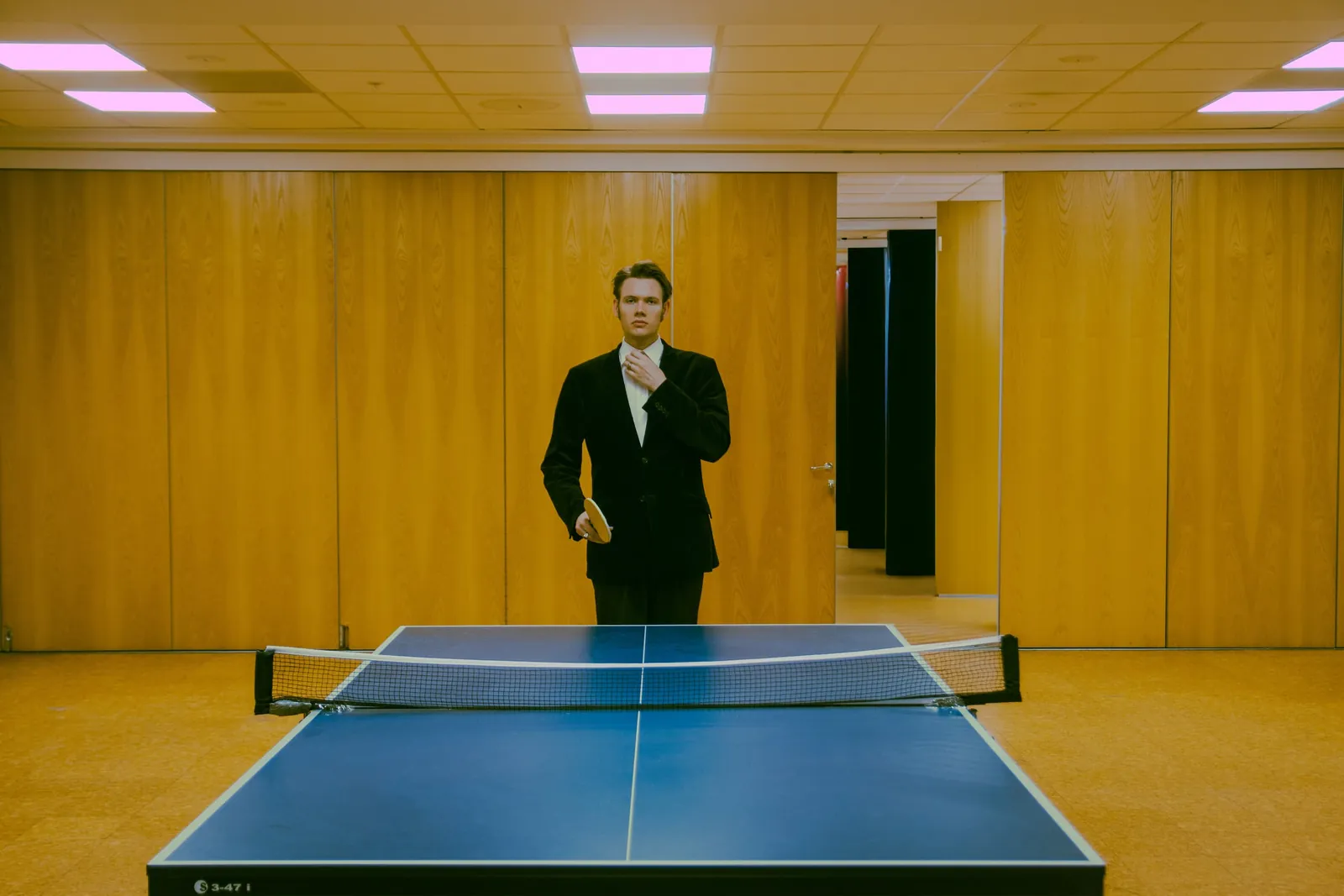 Hubert Kurkiewicz in a suit standing behind a table tennis table in a yellow-panelled hall.
