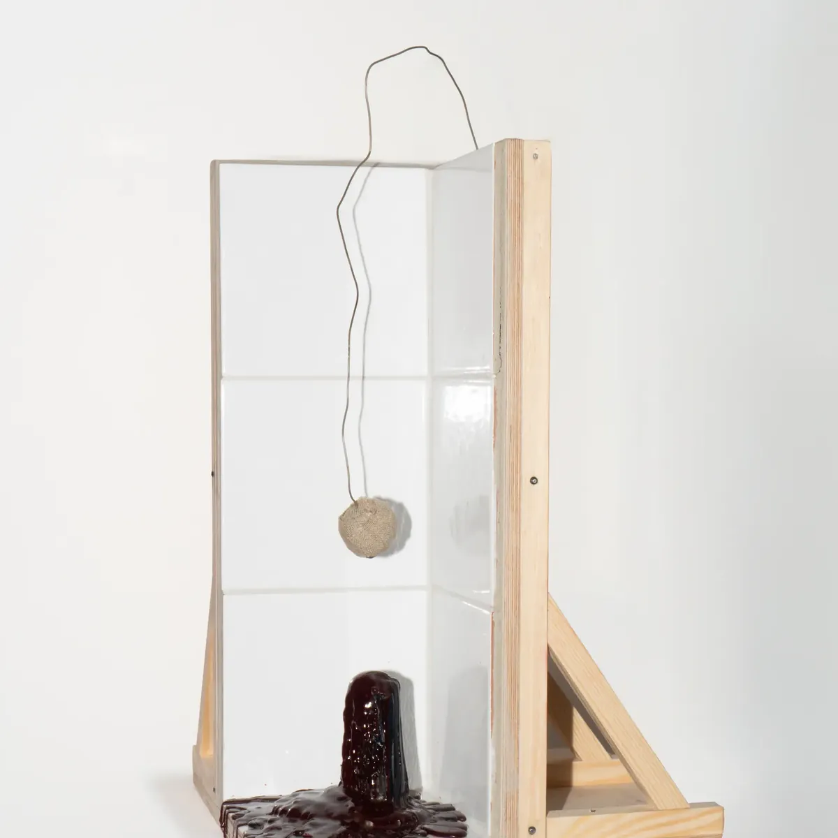 A plywood frame holding a thin wire from which a small clay weight hangs above a dark red base.
