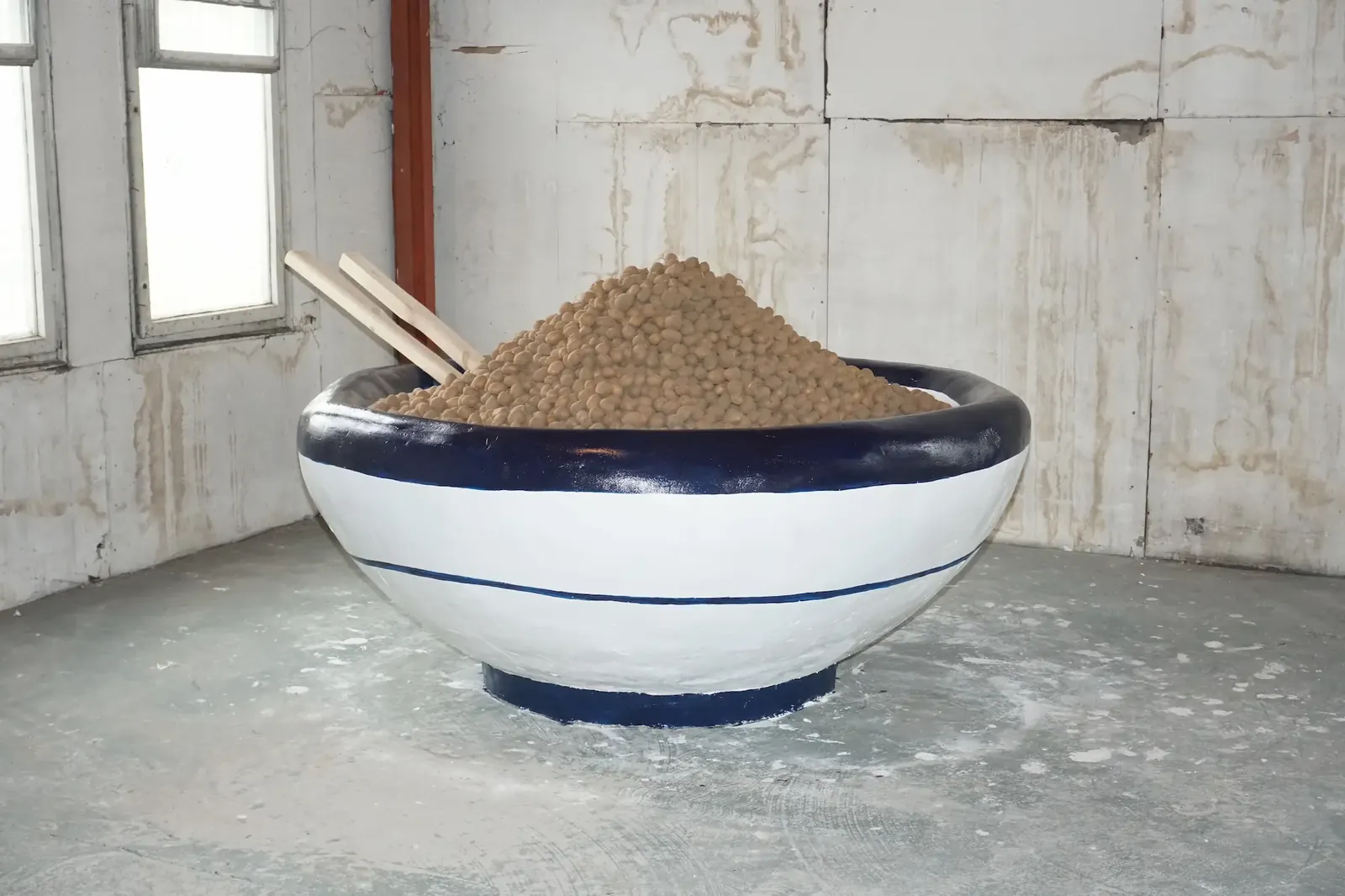 An oversized white and blue ceramic bowl heaped with potatoes, standing in a bare concrete room.