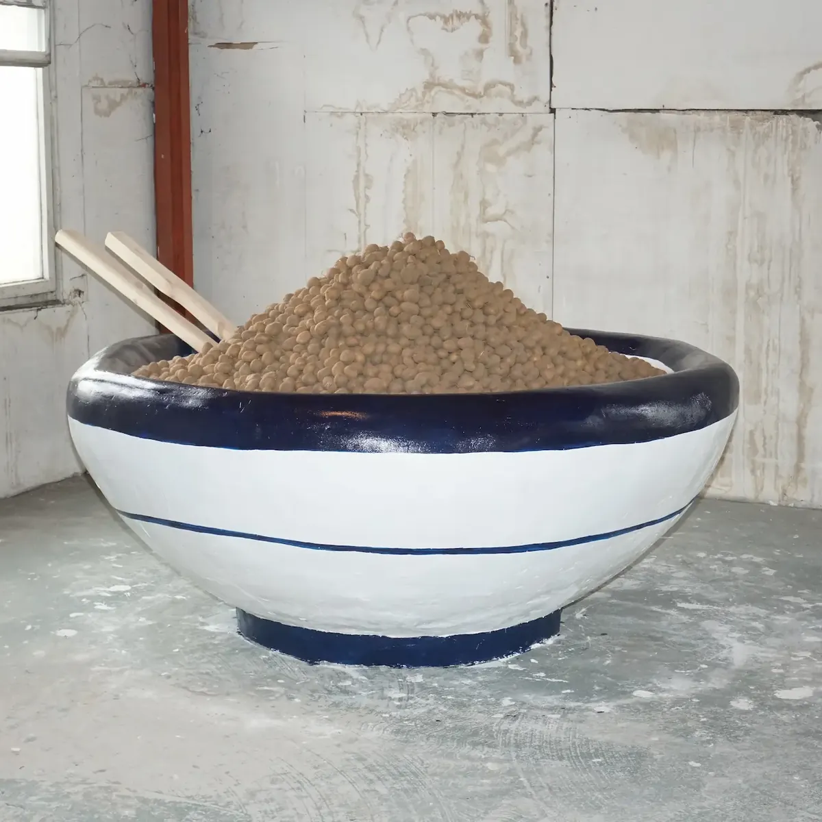 An oversized white and blue ceramic bowl heaped with potatoes, standing in a bare concrete room.