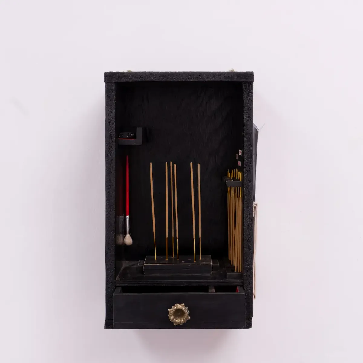 A small black wooden shrine mounted on a wall, holding incense sticks, a brush and a lighter.