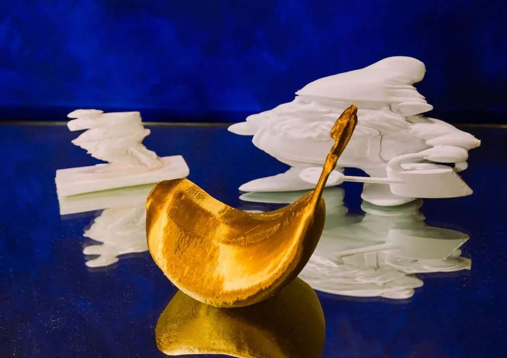 A golden distorted fruit form beside white 3D-printed shapes on a deep blue ground.