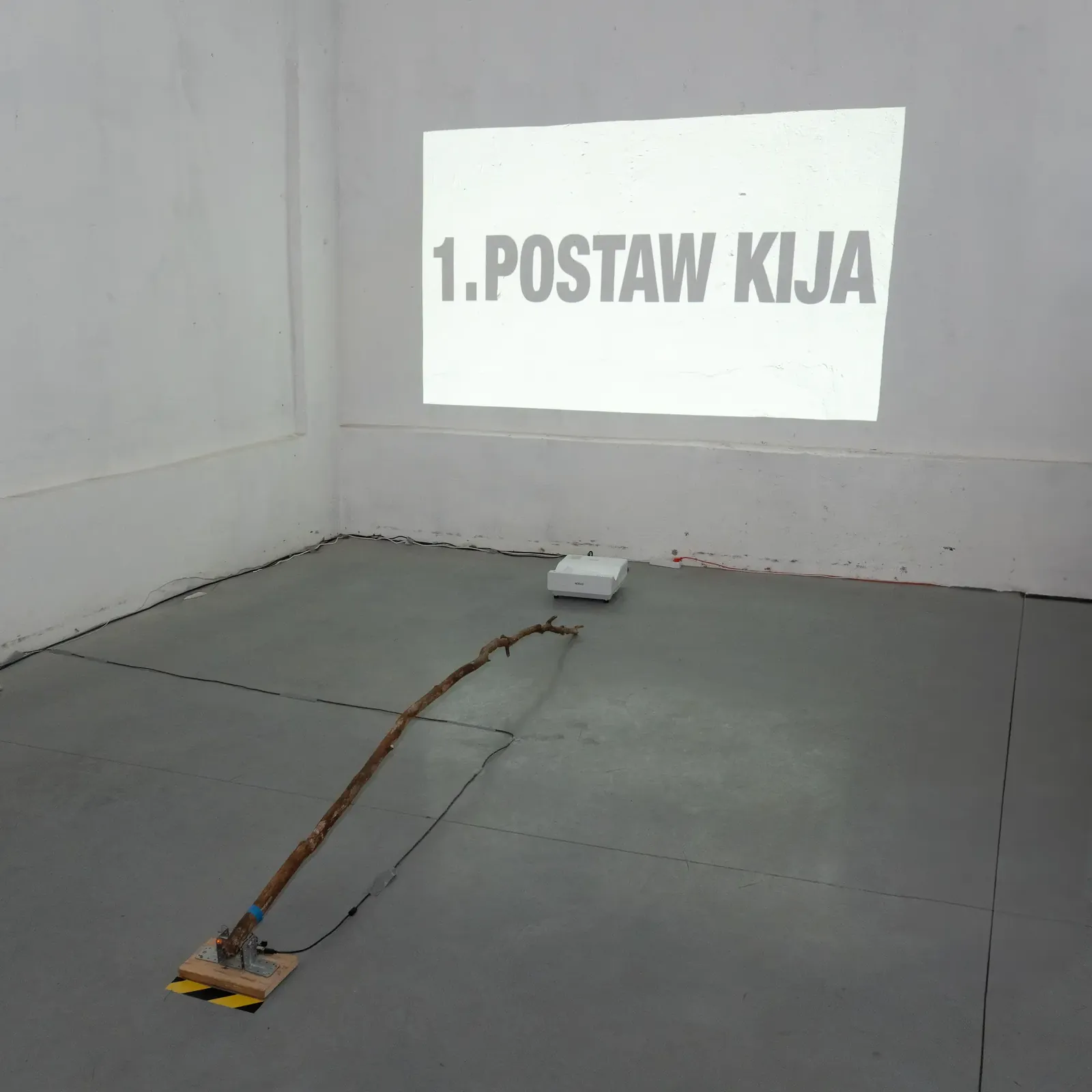 A projection reading “1. POSTAW KIJA” on a gallery wall, with a long wooden stick lying on the floor below.