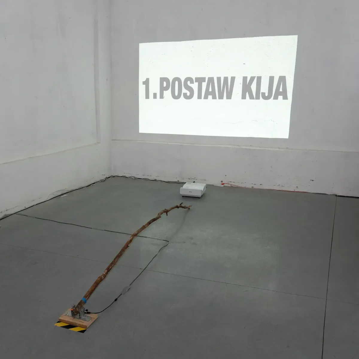 A projection reading “1. POSTAW KIJA” on a gallery wall, with a long wooden stick lying on the floor below.