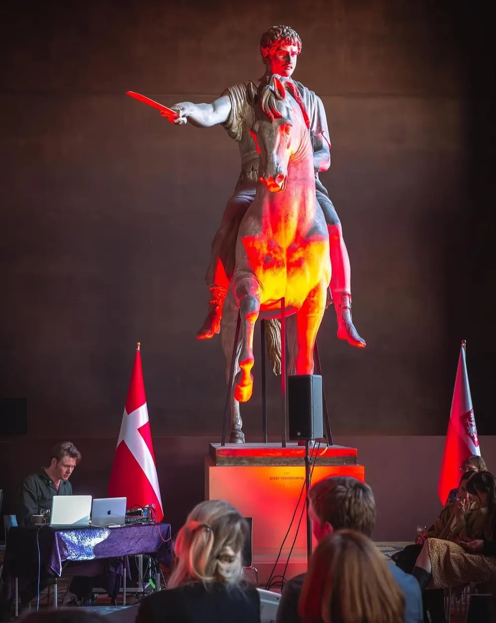 A statue lit in red with projected light, an audience seated in front of it.