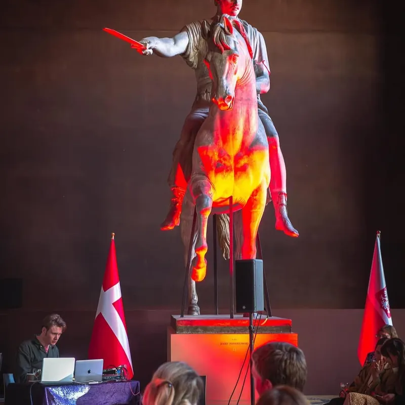 A statue lit in red with projected light, an audience seated in front of it.