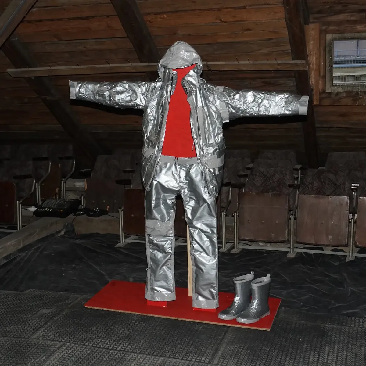A work suit entirely wrapped in silver duct tape, standing upright in an attic space on a red mat.